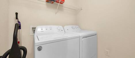 Un espace buanderie avec un lave-linge et un sèche-linge blancs, un aspirateur et du détergent rangés sur une étagère.