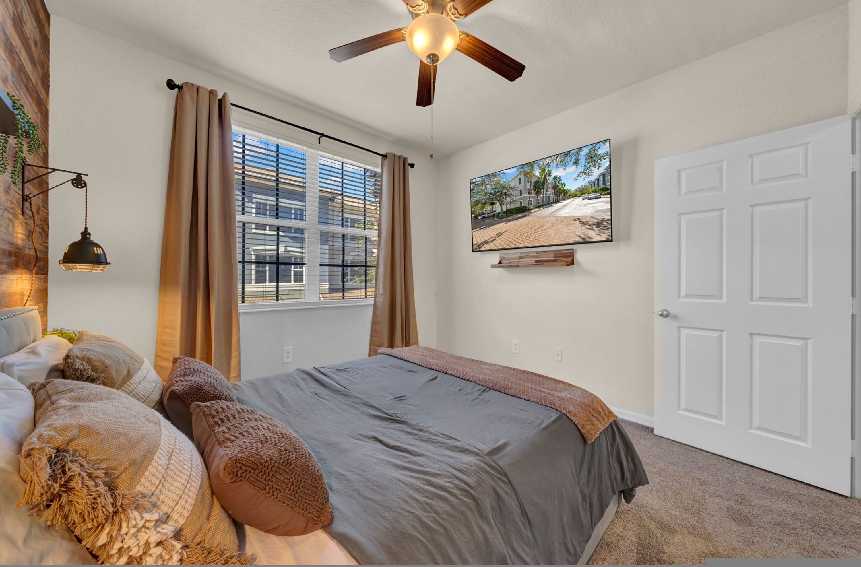 A bedroom with a bed, a ceiling fan, a window with curtains, a mounted TV, and a door.