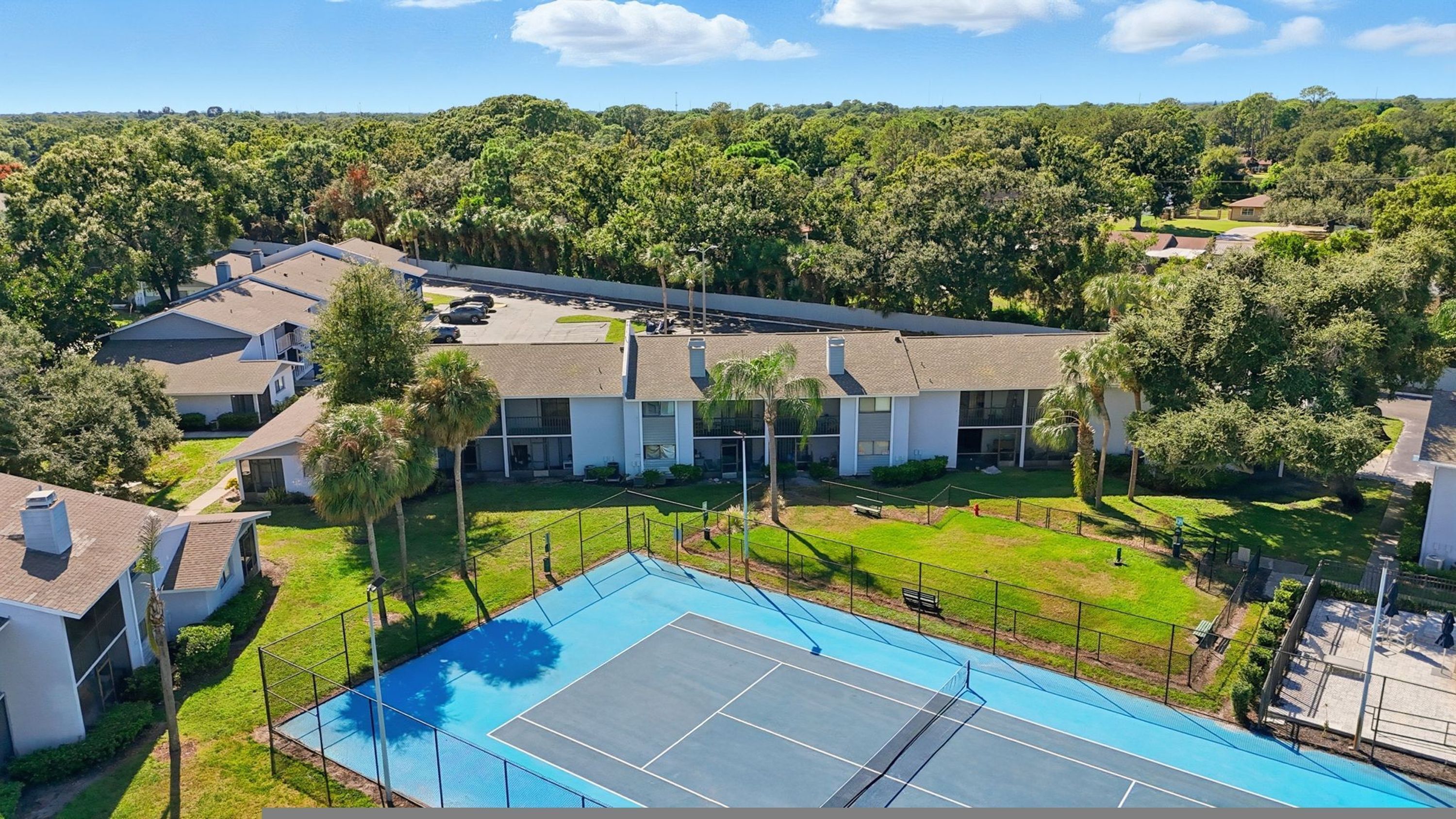 A residential area with a tennis court, houses, and a dense forest in the background.
