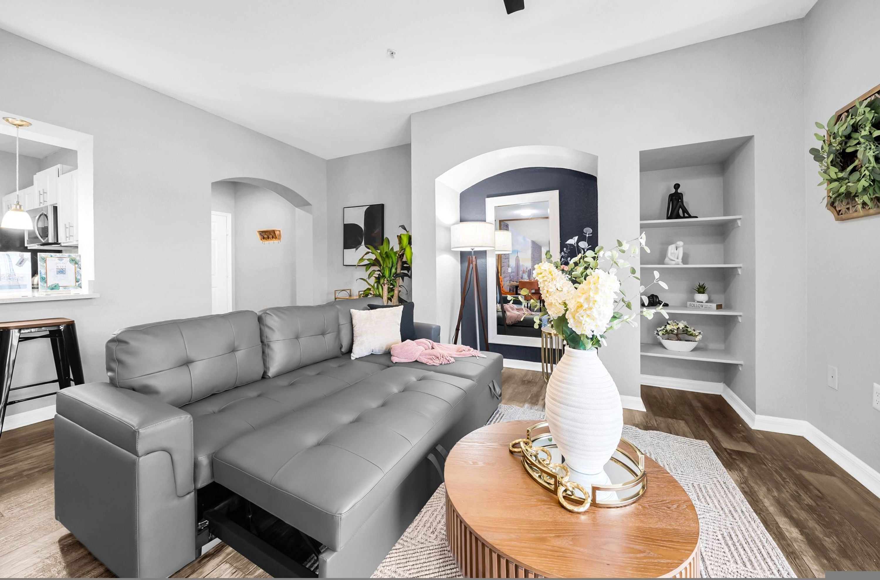 A modern living room with a grey sofa, a round wooden coffee table with a vase, and a kitchen area visible through an archway.