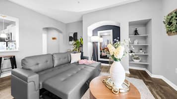 A modern living room with a grey sofa, a round wooden coffee table with a vase, and a kitchen area visible through an archway.