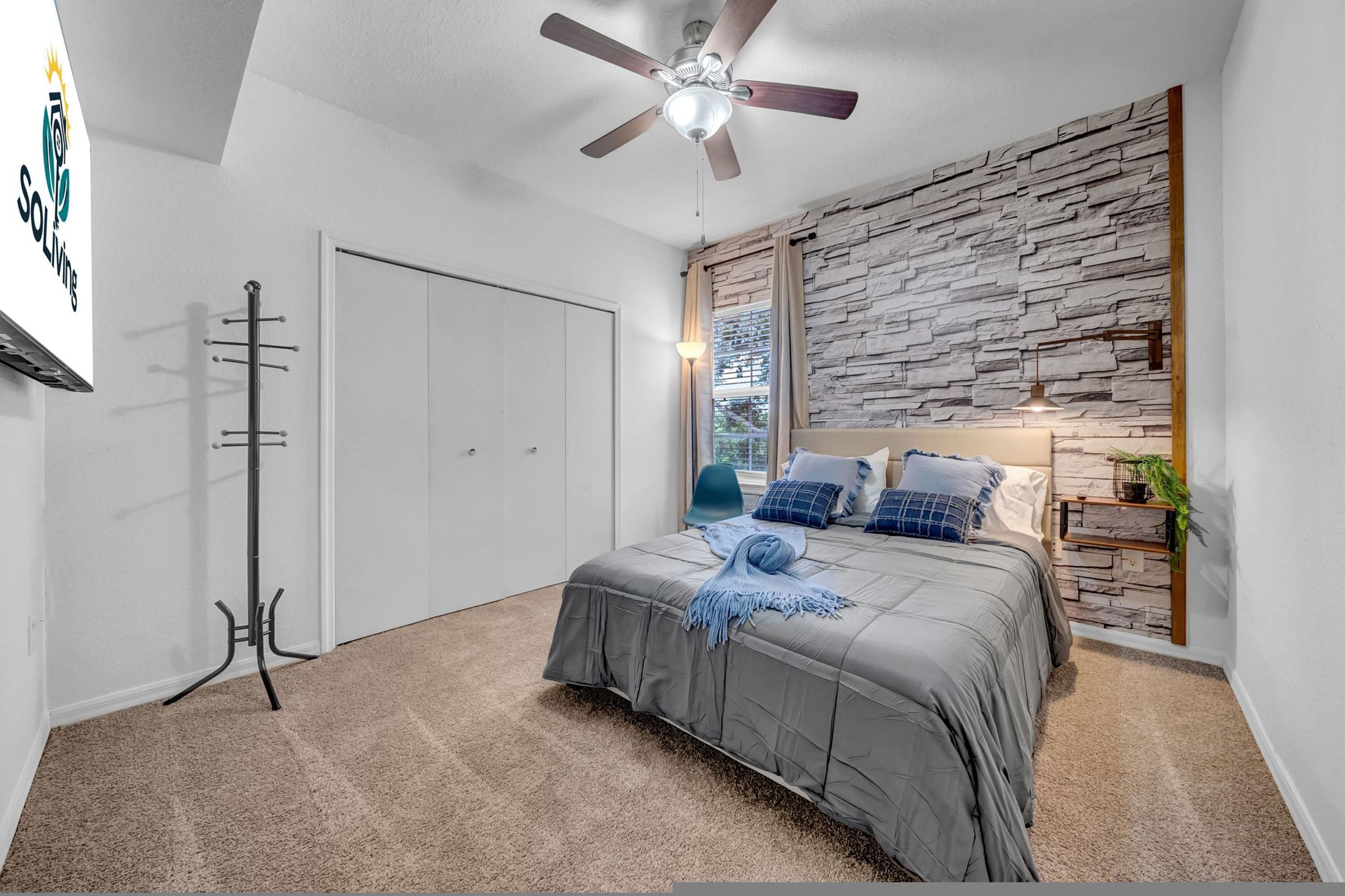 A bedroom with a bed, a ceiling fan, a stone wall, a window, and a nightstand with a lamp.