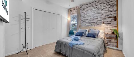 A bedroom with a bed, a ceiling fan, a stone wall, a window, and a nightstand with a lamp.