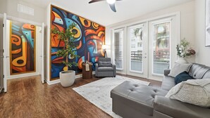 A modern living room with hardwood flooring, a grey sofa, a grey armchair, a large abstract painting, and a ceiling fan. - Stylish 2BR w Pool Pet-friendly Unit 13 (Tampa)