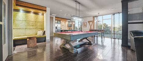 A modern indoor pool table with a wooden frame, a seating area with a patterned cushion, and a large window providing a view of the outdoors.