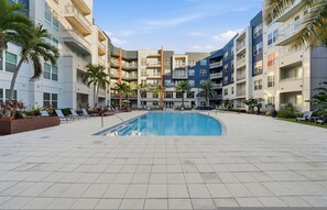 A swimming pool surrounded by multi-story buildings with balconies, palm trees, and lounge chairs. - Stylish 2BR w Pool Pet-friendly Unit 13 (Tampa)