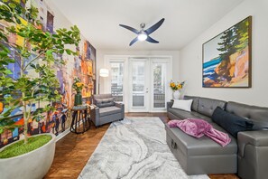 A living room with a gray sofa, a coffee table, a ceiling fan, a large abstract painting, and a potted plant. - Chic 2BR w Pool Pet-friendly Unit 14 (Tampa)