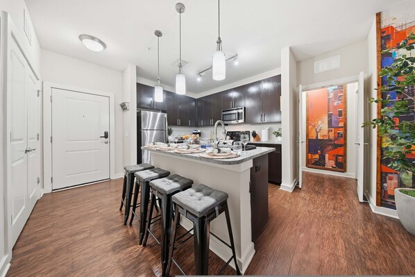 A modern kitchen with dark cabinets, a central island with stools, stainless steel appliances, pendant lighting, and a cityscape painting. - Chic 2BR w Pool Pet-friendly Unit 14 (Tampa)