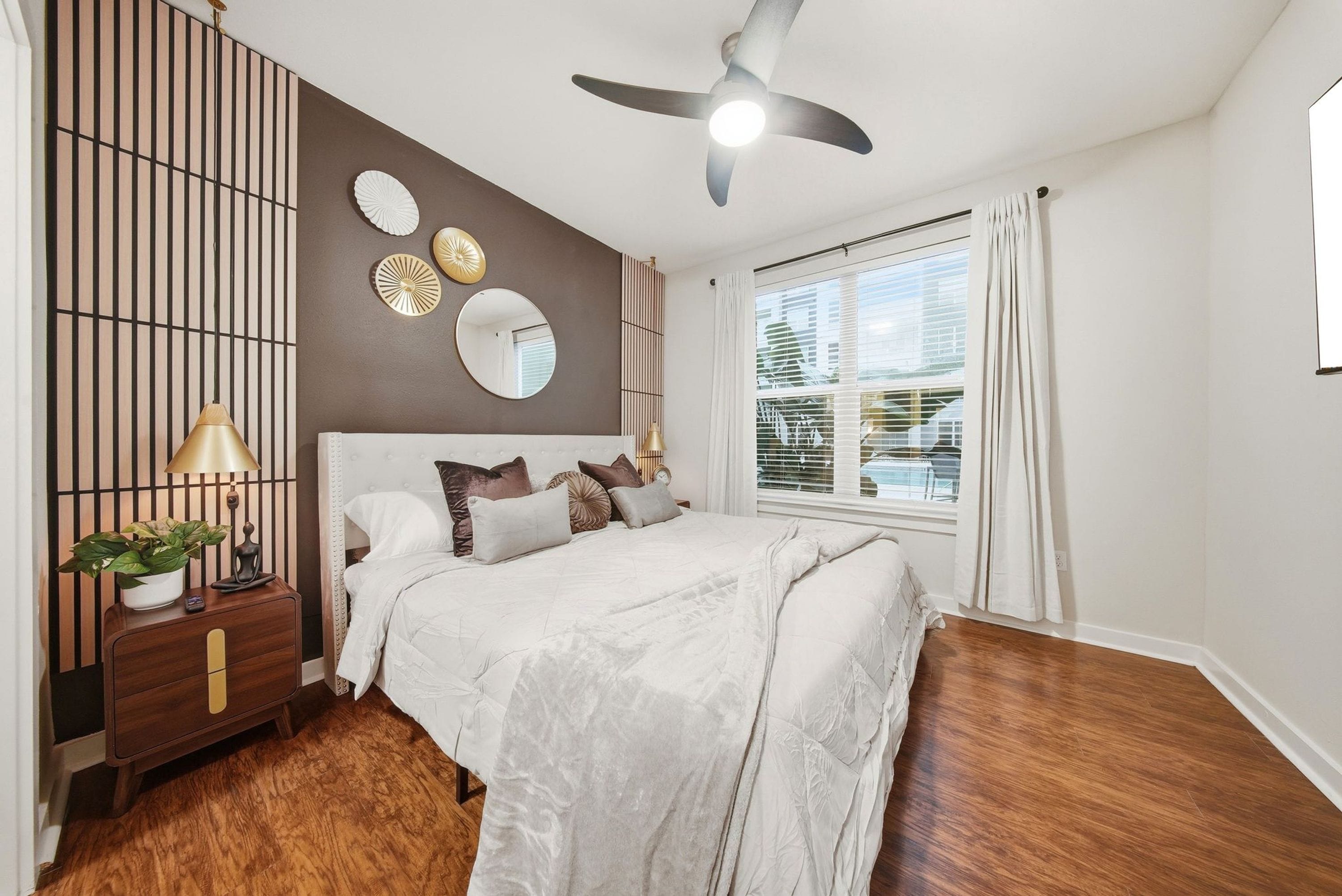 Une chambre à coucher comprenant un lit, une table de chevet, un ventilateur de plafond, une fenêtre avec des rideaux et un mur décoré d’assiettes.
