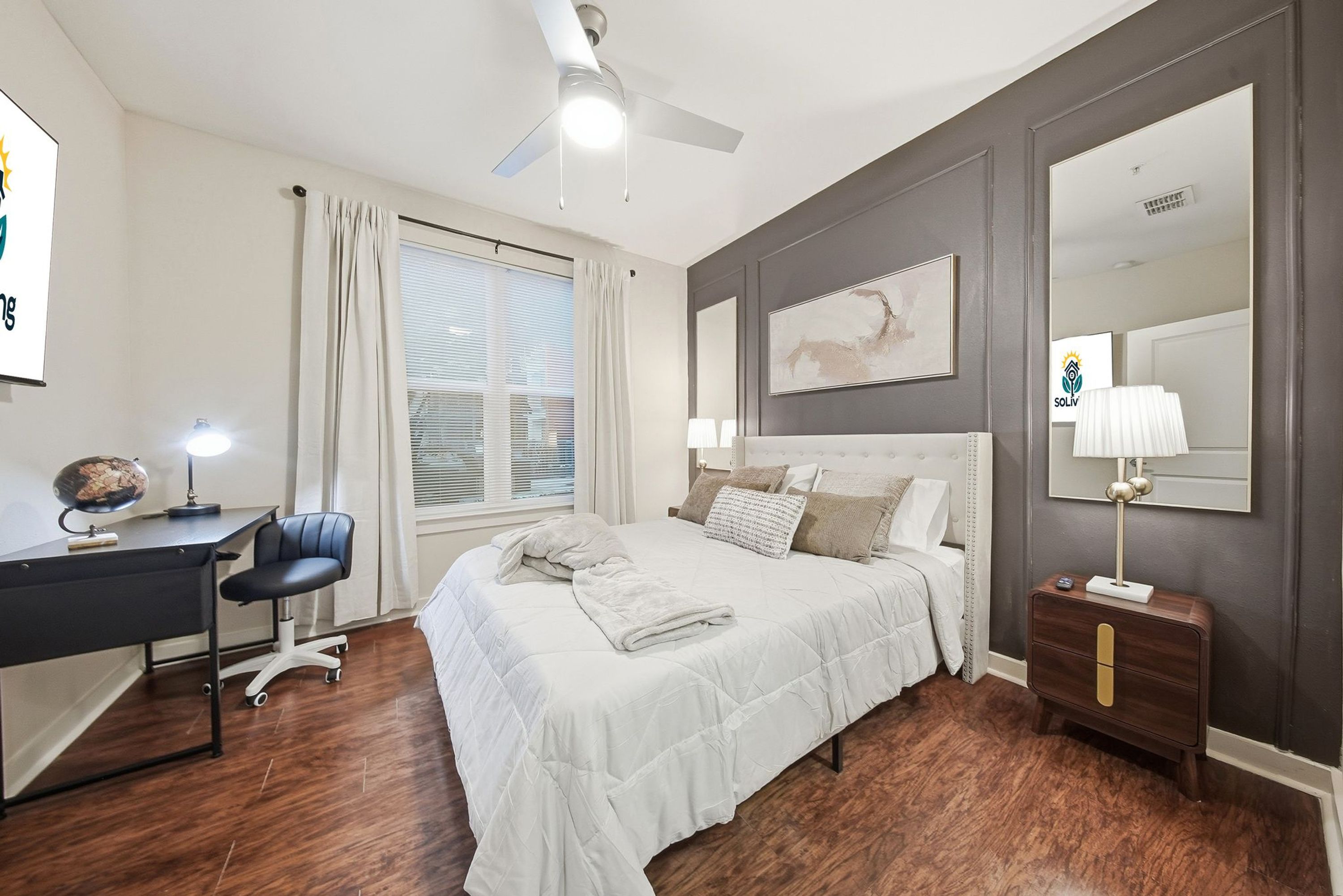 A bedroom with a bed, desk, chair, nightstand, lamp, and a ceiling fan.