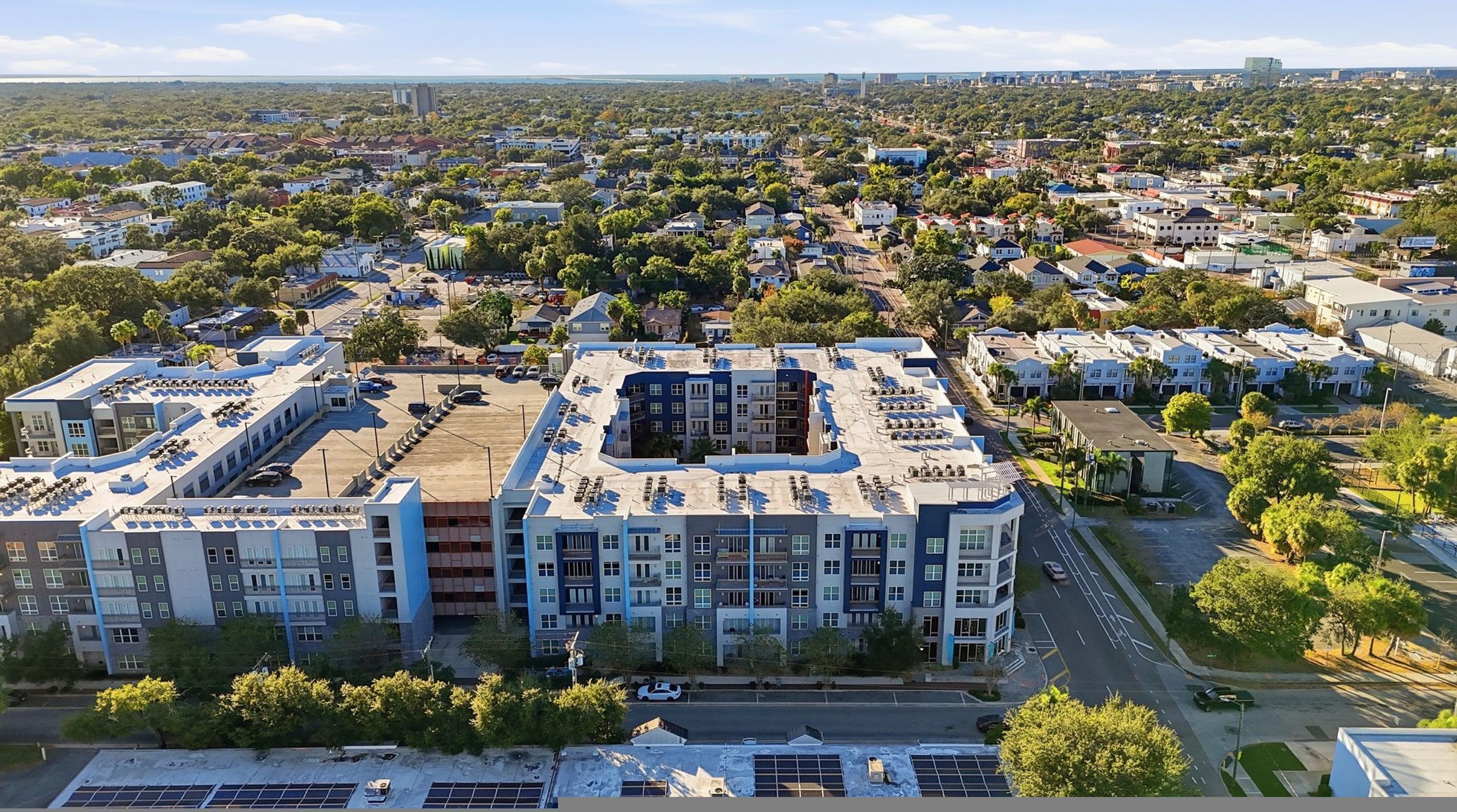 A multi-story residential building with a parking lot, surrounded by greenery and other residential buildings.