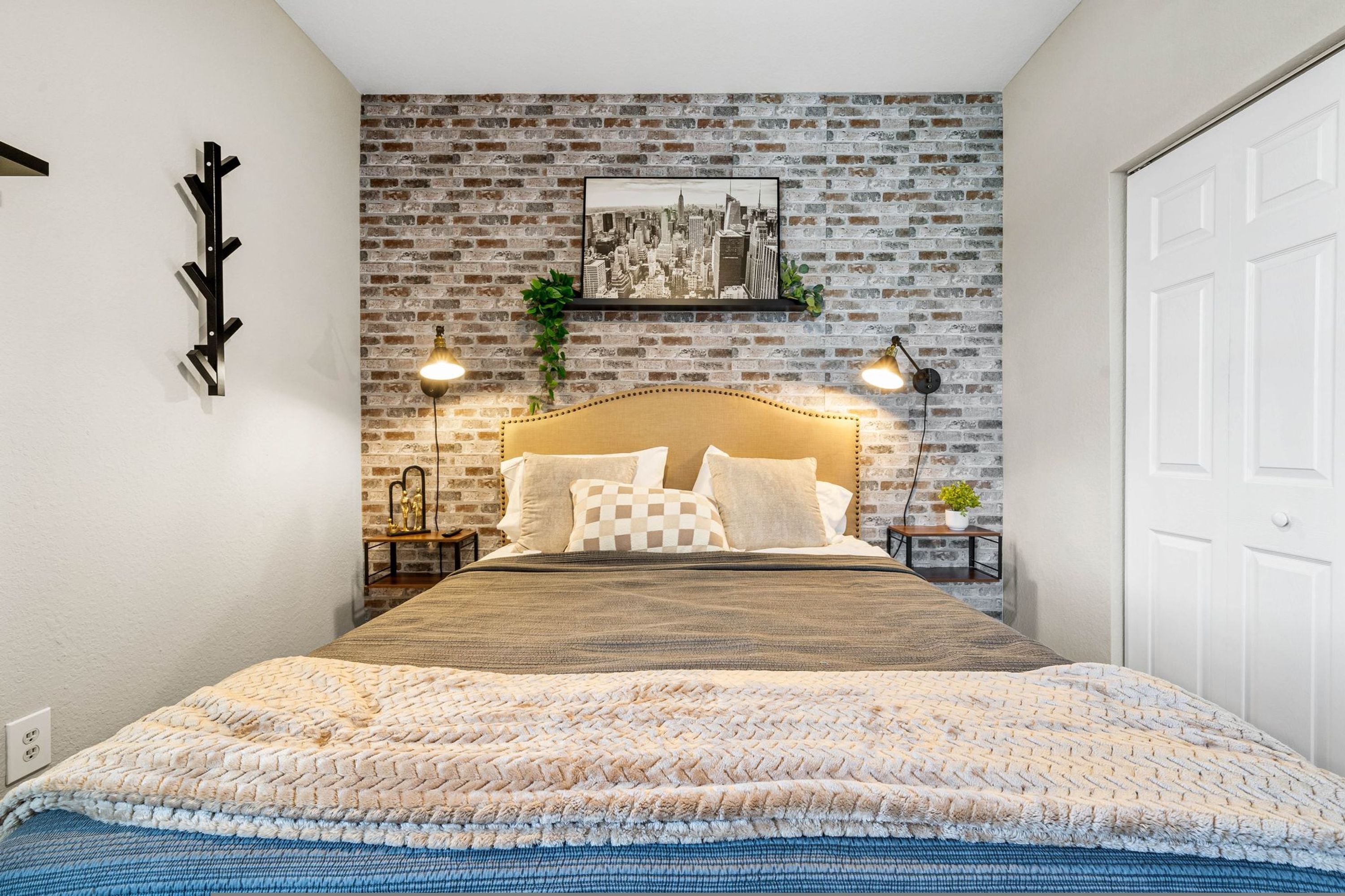 A bedroom with a brick-patterned wall, a cityscape picture, a bed with a headboard, bedside lamps, and a coat rack.