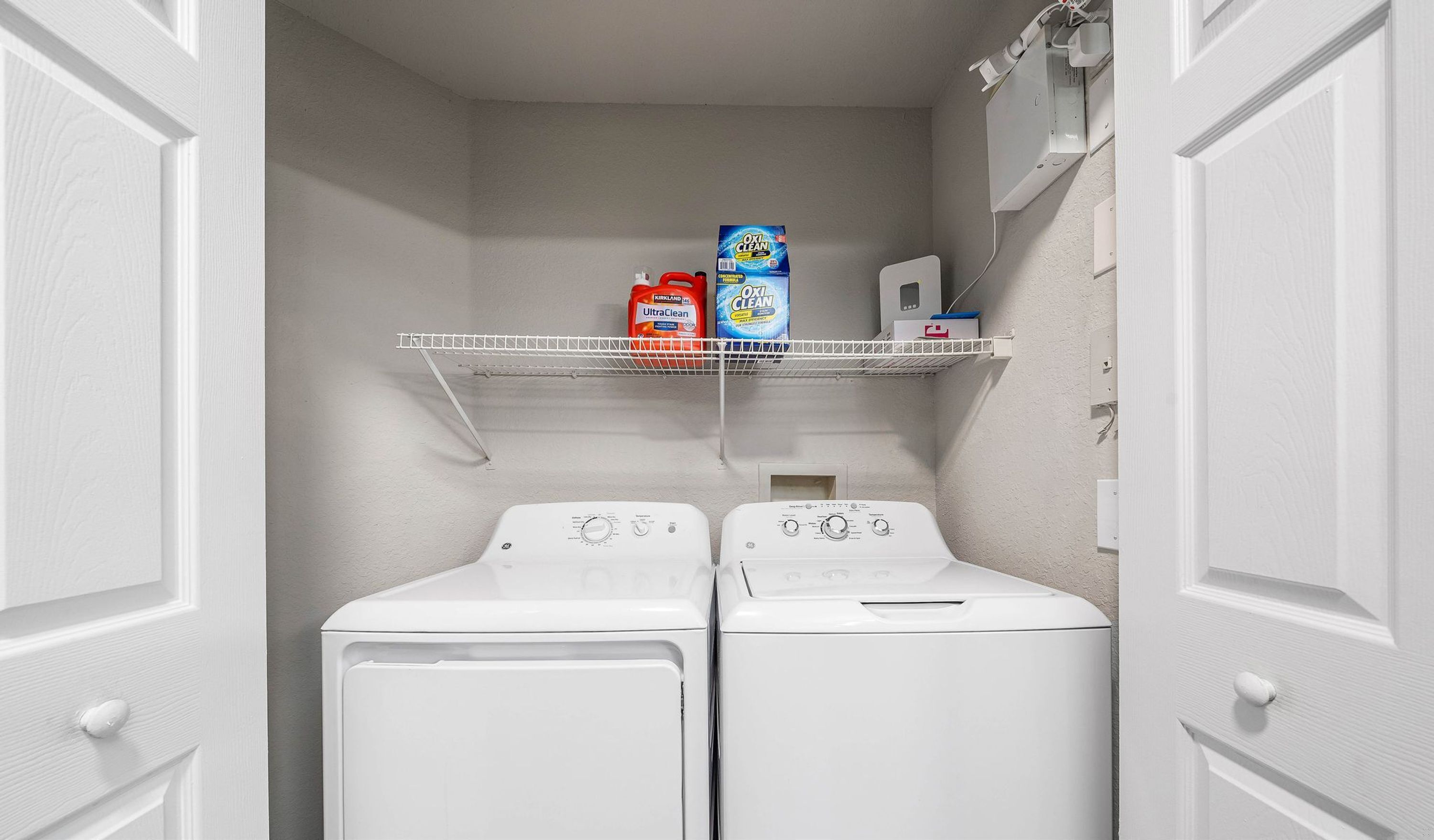 A laundry room with two front-loading washing machines, a shelf with detergent bottles, and a control panel.