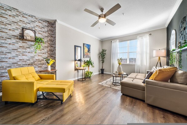 A living room with a brick wall, a ceiling fan, a yellow sofa, a beige sofa, and a wooden floor. - Modern 3BR 2BA Pools Gym Near Airport 5 (Tampa)