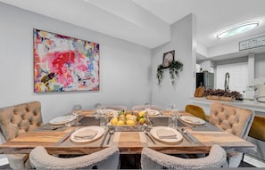 Uma sala de jantar com uma mesa de madeira posta para seis pessoas, um quadro abstrato colorido na parede e uma área de cozinha visível ao fundo.