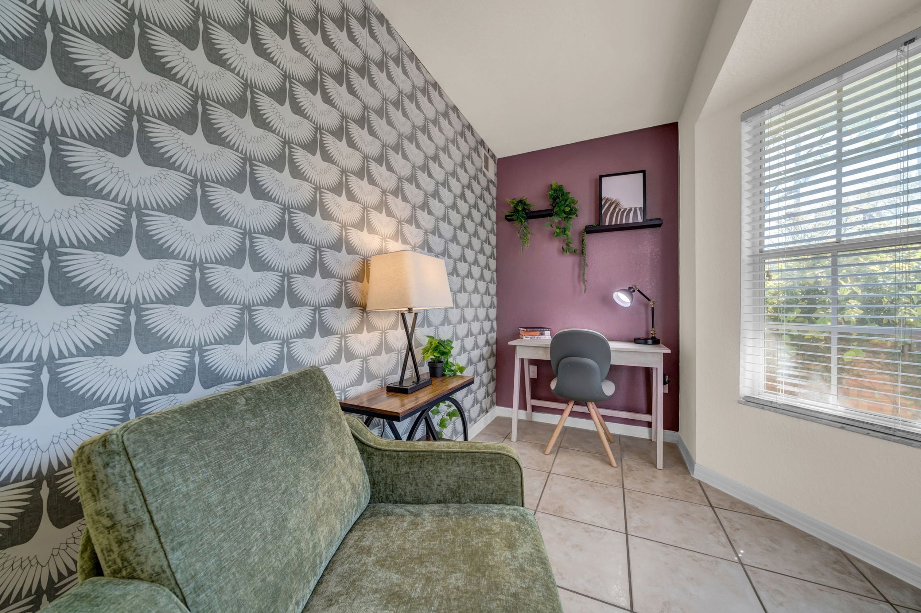A room with a patterned wallpaper, a green sofa, a desk with a lamp, and a window with blinds.