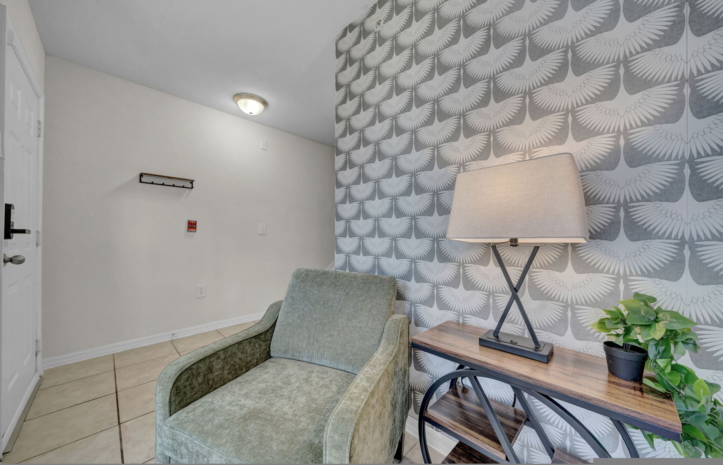 A room with a patterned wallpaper, a chair, a table with a lamp, and a potted plant.