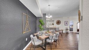 Un coin repas avec une table en bois dressée pour quatre personnes, des chaises à l’assise rembourrée de couleur grise, un mur à rayures noires et blanches, et un grand tableau encadré représentant la Tour Eiffel.