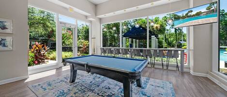 A pool table in a spacious room with large windows, a view of a pool, and a gazebo outside.