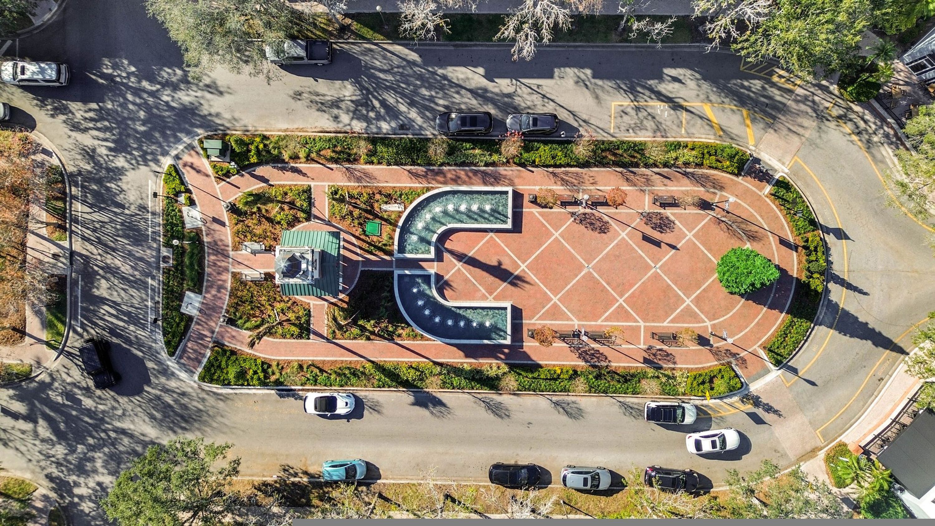 Vista aérea de un estacionamiento con un amplio espacio verde de forma singular, rodeado de árboles y una carretera.