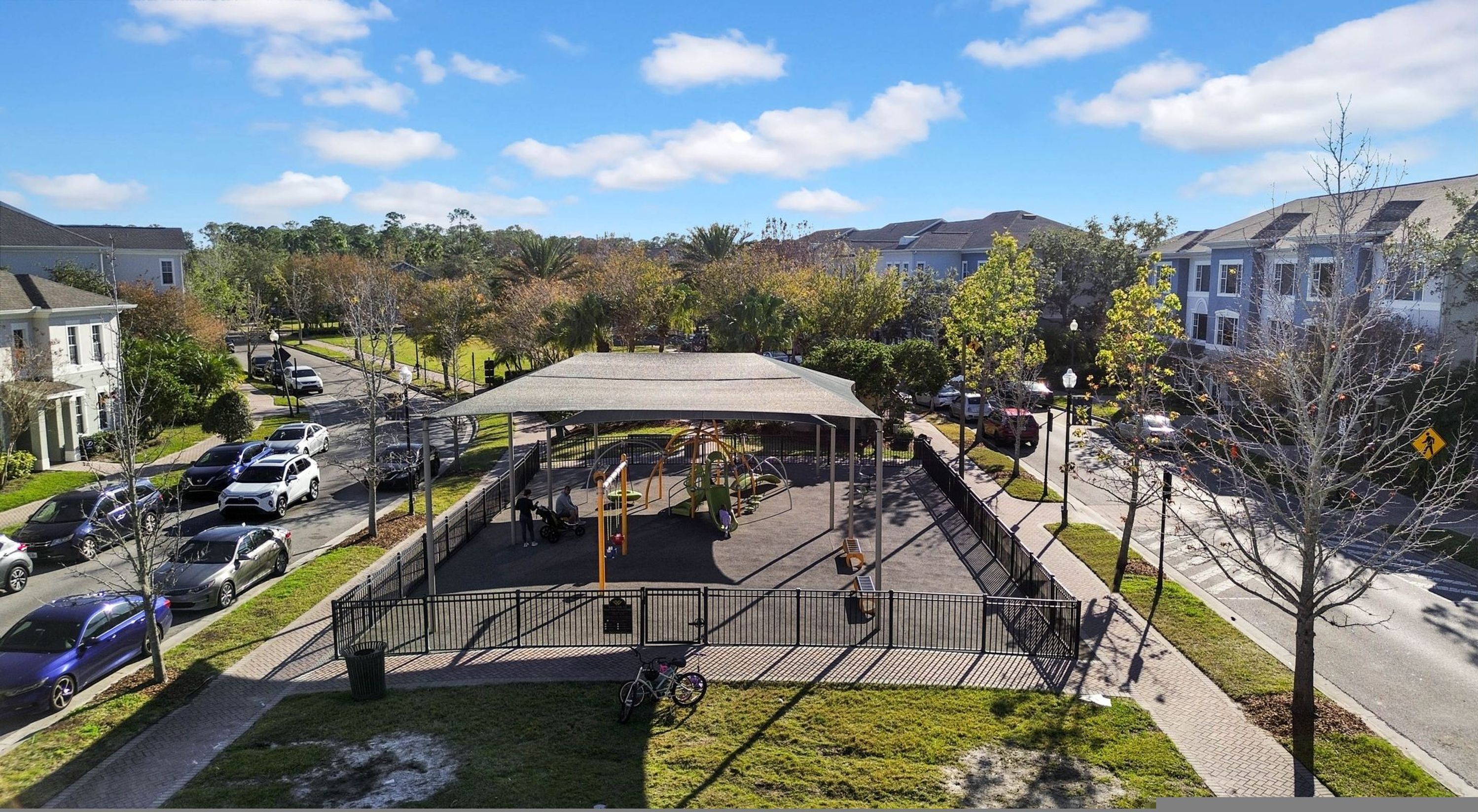 Un parque infantil con zona de juegos cubierta, rodeado de coches aparcados y edificios residenciales.
