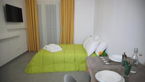 A hotel room with a green bed, a dining table with plates and cutlery, a chair, and a TV mounted on the wall. - Farina Rooms & Apartments - Hospitality with a View and Comfort in the City (San Nicola La Strada)