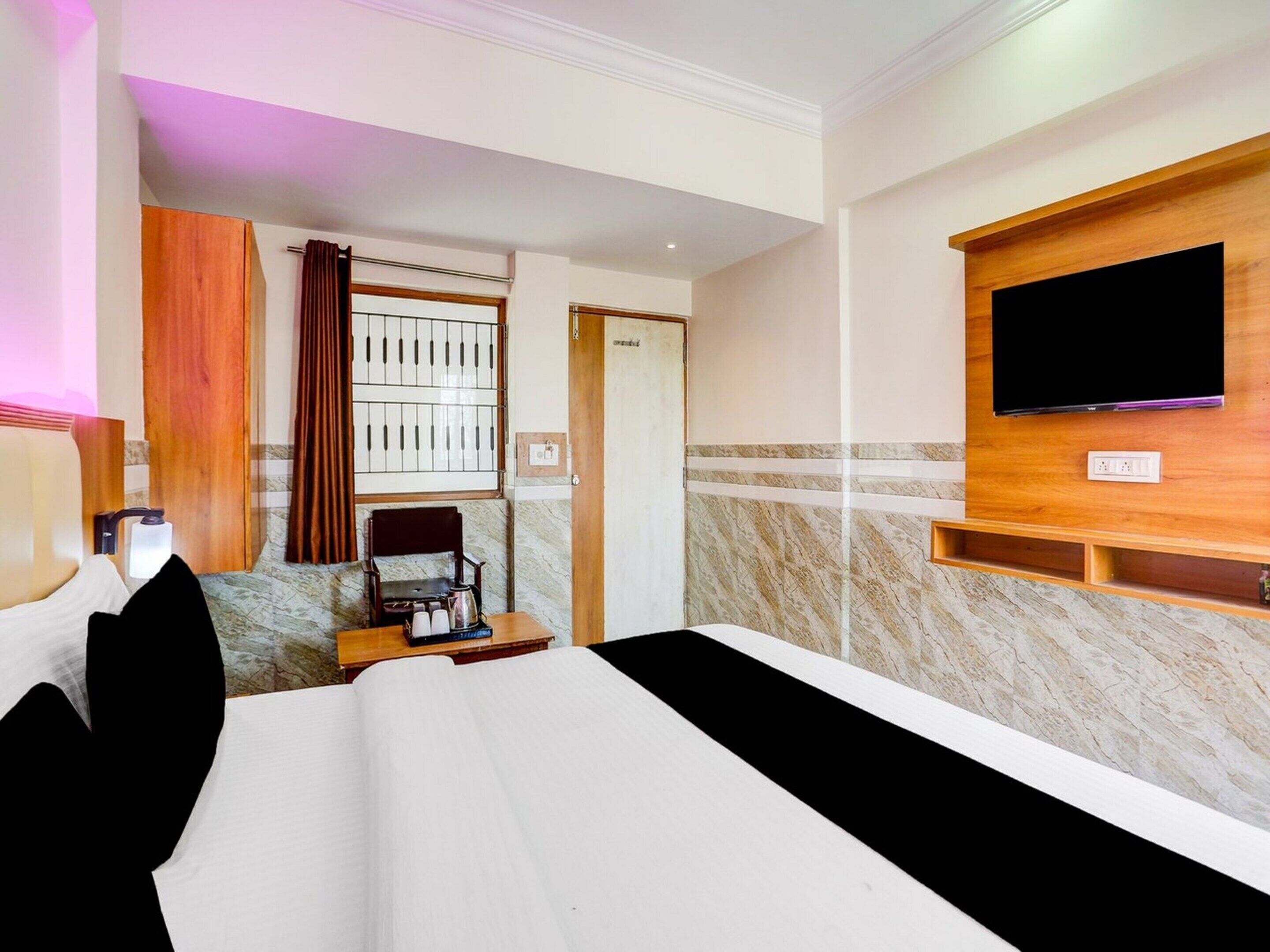 A hotel room with a bed, a TV mounted on the wall, a desk with a chair, a window with curtains, and a door.
