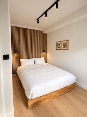 A modern bedroom with a wooden bed frame, a slatted wall panel, and two wall-mounted lights. - Executive 1 Bedroom Apartment (Apt C) (London)
