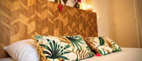 Una cama con ropa de cama blanca y dos almohadas con estampado de hojas tropicales, un cabecero de madera, espejos montados en la pared y un florero con flores.