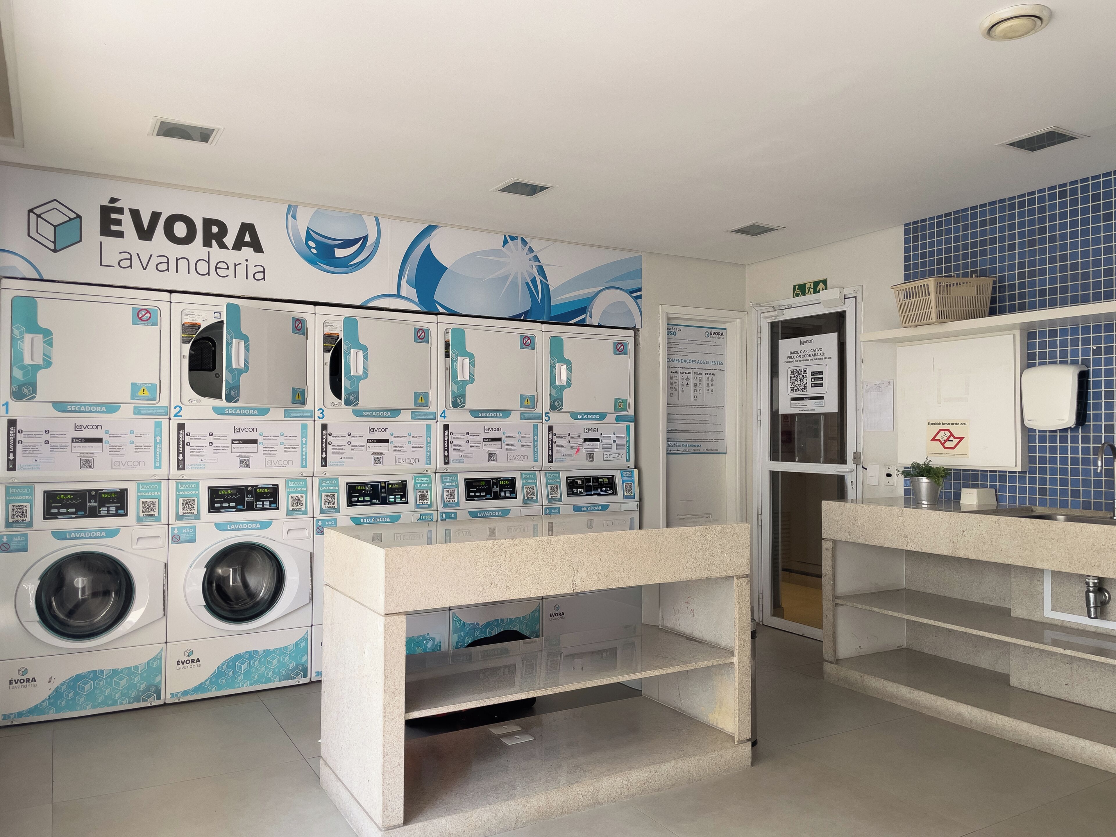 A laundromat with multiple washing machines and dryers, a sink area, and a blue tiled wall.