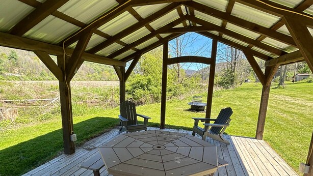 Eine überdachte Terrasse im Freien mit einem sechseckigen Tisch und Picknickbänken, umgeben von einer Holzterrasse und Blick auf ein grünes Feld.