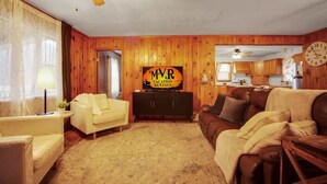Ein Wohnzimmer mit einer Holzvertäfelung, einem Deckenventilator, einem Fernseher, einem Sofa, einem Couchtisch und einer Küche im Hintergrund.