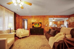 A living room with a wooden wall, a ceiling fan, a TV, a sofa, a coffee table, and a kitchen in the background. - Misty Mountain Cottage - Pet Friendly (Shippen Township)