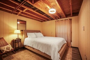A bedroom with a wooden ceiling, a bed with white bedding, a nightstand with a lamp, and a chair. - Misty Mountain Cottage - Pet Friendly (Shippen Township)