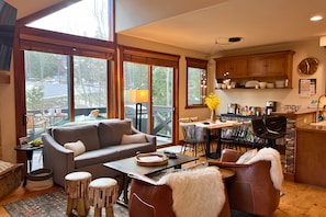 A spacious living area with a sofa, coffee table, and dining table. Large windows offer a view of the outdoors. - Trailhead Basecamp • Ski/Bike‑In/Out • Hot Tub (Breckenridge)