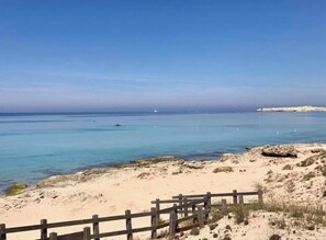 Uma praia com água cristalina, uma cerca de madeira e barcos ao longe.