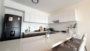 A modern kitchen with white cabinets, a black refrigerator, a marble countertop, and a small island with chairs. - Luxurious brand-new apartment just a few blocks from Larcomar (Miraflores)