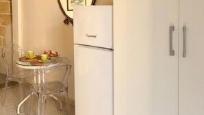 Une cuisine avec un réfrigérateur blanc, des chaises transparentes, une petite table avec de la nourriture et un tableau encadré au mur.
