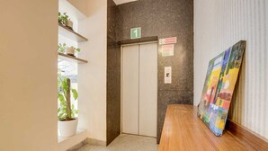 Un pasillo con un banco de madera, un cuadro, una planta en maceta, un ascensor y un cartel de 'No Fumar'.