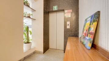 A hallway with a wooden bench, a painting, a potted plant, an elevator, and a 'No Smoking' sign.