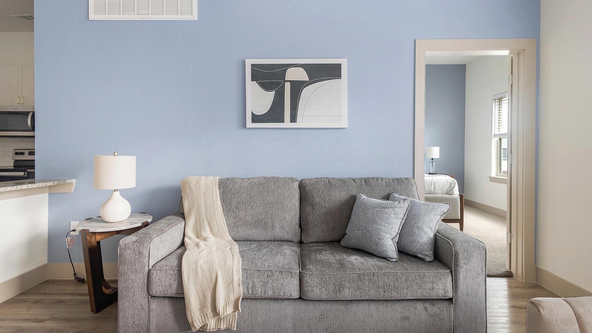 A living room with a grey sofa, a white lamp, and a framed artwork on the wall.