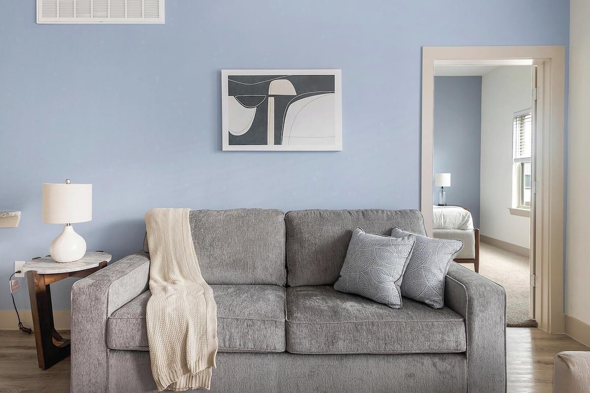 A living room with a grey sofa, a white lamp, a framed abstract artwork, and a kitchen area in the background.