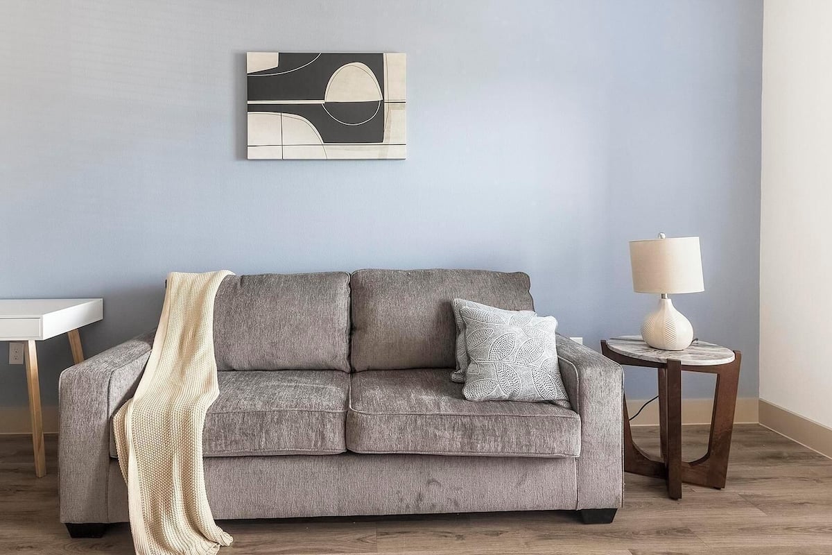 A grey sofa with a throw blanket and patterned pillows, a white desk, a small round table with a lamp, and abstract wall art.