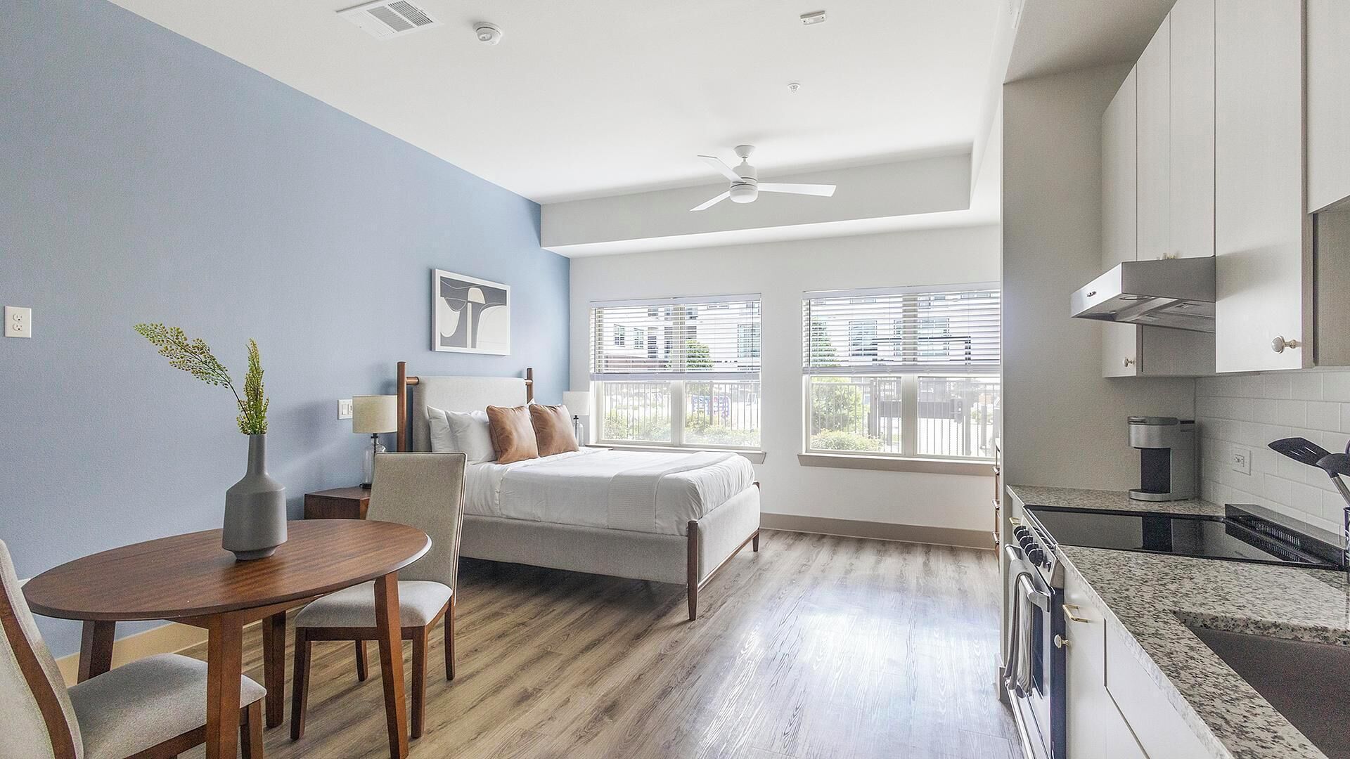 Una habitación de hotel moderna con cama, zona de comedor, cocina y amplias ventanas.
