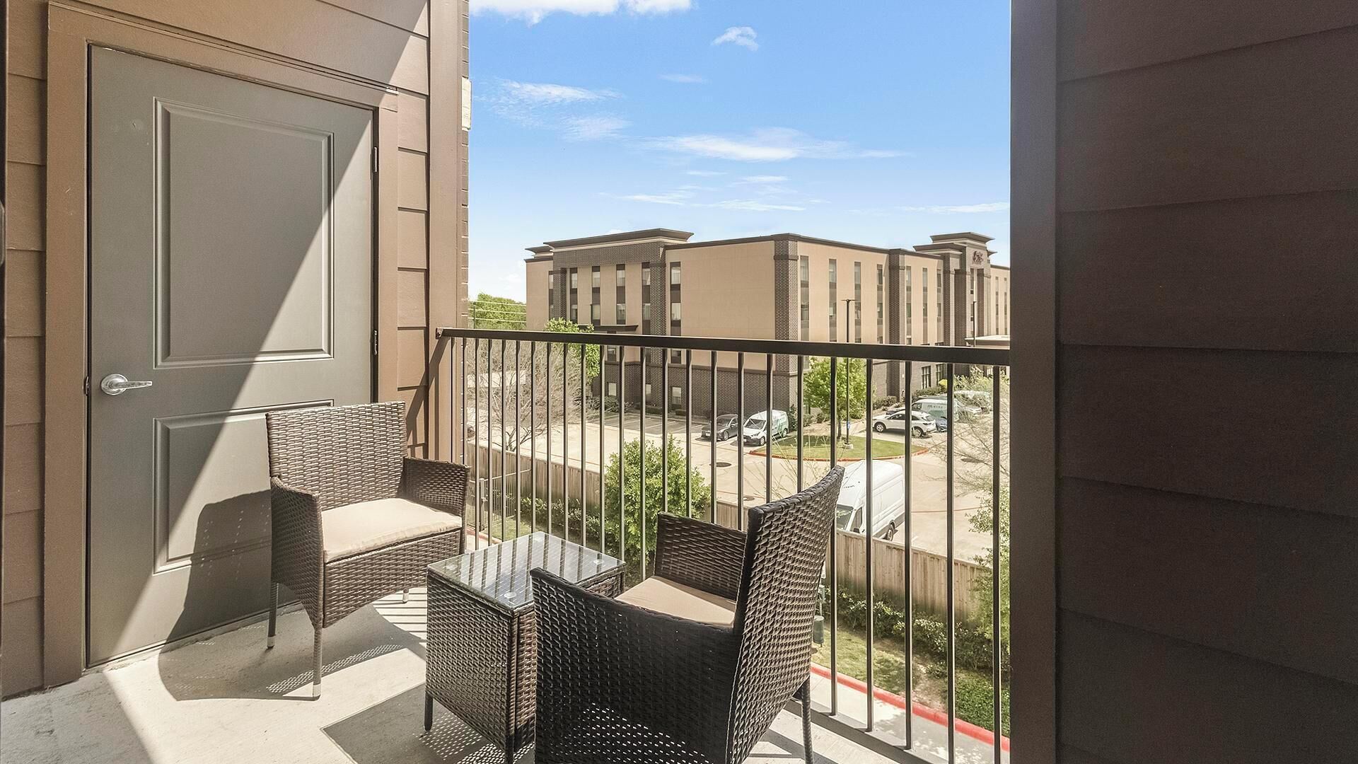 A balcony with wicker furniture, a glass table, and a view of buildings and parked cars.