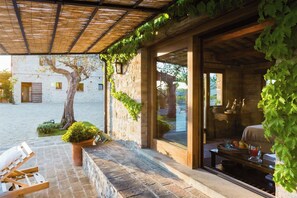 Un espace extérieur aménagé, abrité, avec une table et des chaises, un pot de fleurs et une vue sur un bâtiment doté d’un mur en pierre et d’un arbre.