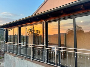 Um edifício moderno com grandes janelas refletindo um pôr do sol, varanda com guarda-corpo de metal e base de concreto.