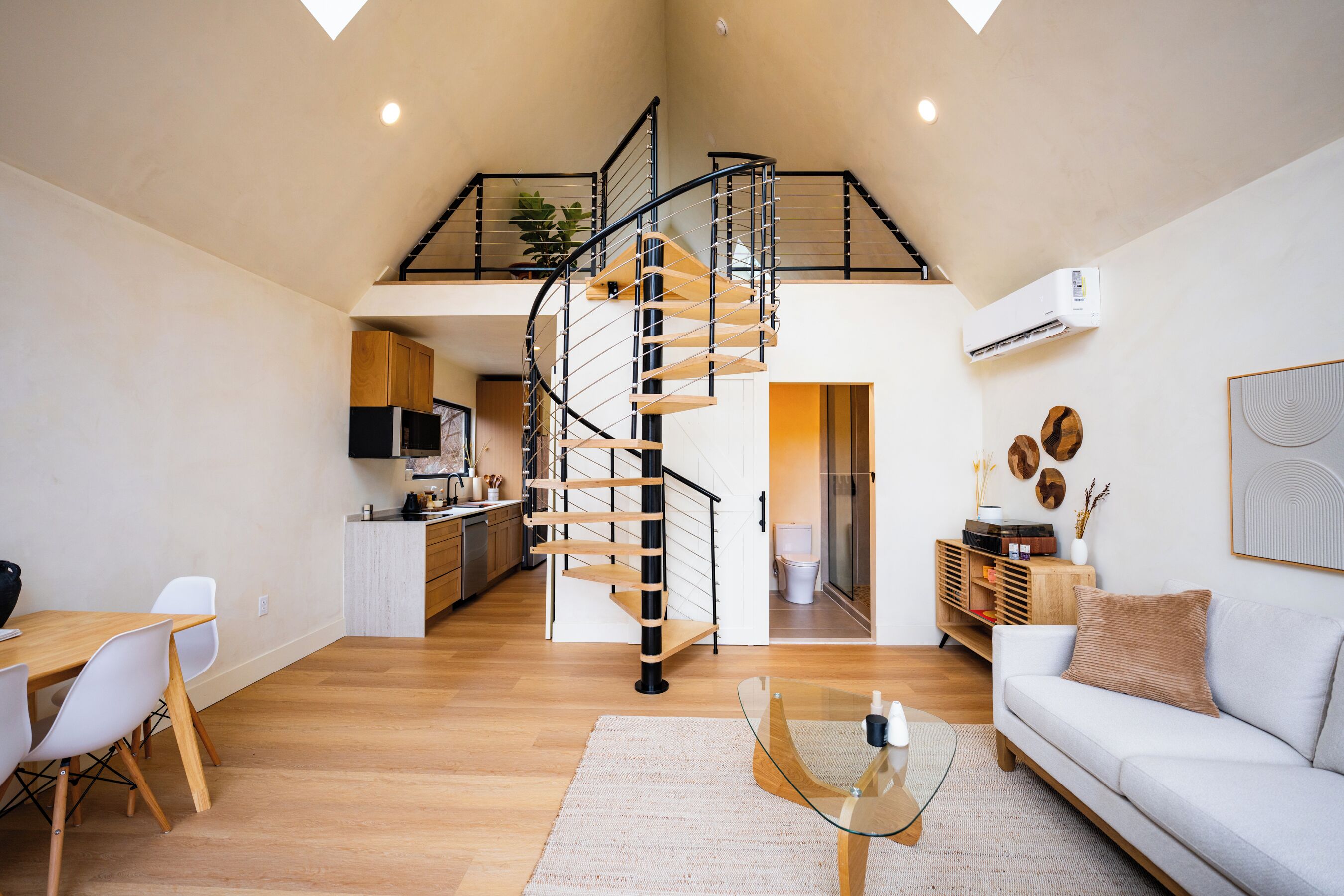 A spacious living area with a spiral staircase, a dining table with chairs, a sofa, and a kitchen in the background.