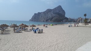 A beach with sun loungers, umbrellas, and a large rock formation in the background. - Apartment 'Rosa De Los Vientos' with Sea View, Wi-Fi, and Air Conditioning (Calpe)