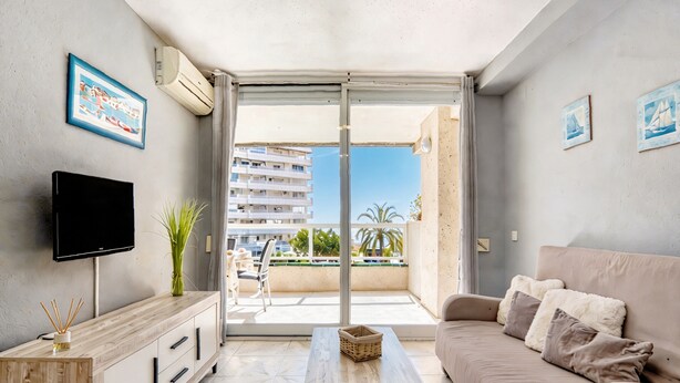 A modern living room with a flat-screen TV, a wooden cabinet, a beige sofa, and a balcony with a view of buildings and palm trees. - Apartment 'Rosa De Los Vientos' with Sea View, Wi-Fi, and Air Conditioning (Calpe)